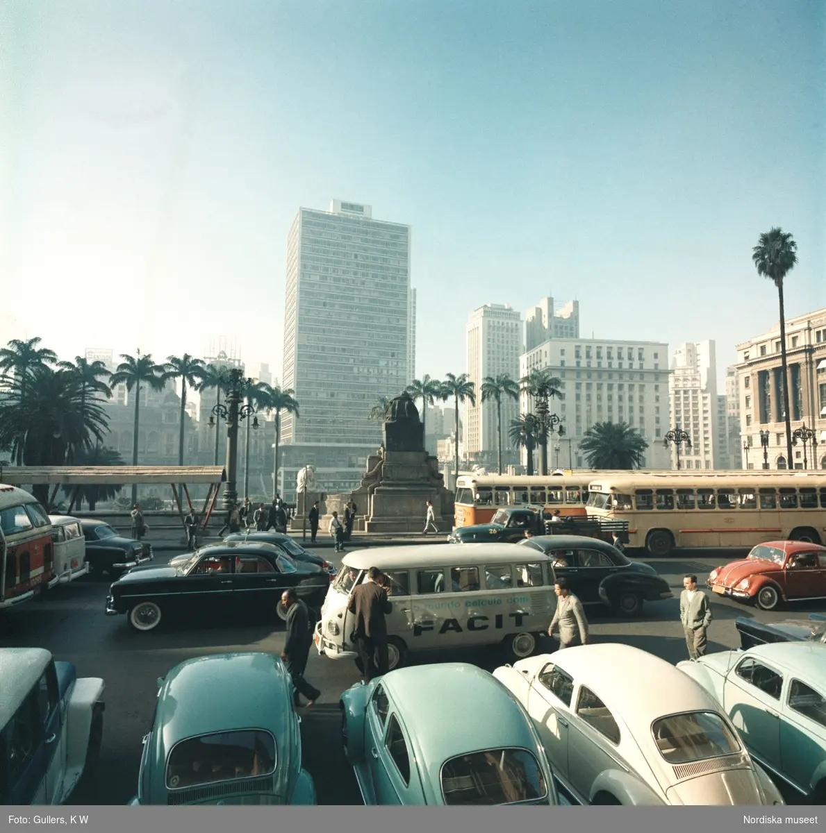 Facit. Brasilien, 1960-tal. Stadsvy med en trafikerad gata i förgrunden. i centrum av bilden en av företagets bilar . En Wolkswagen