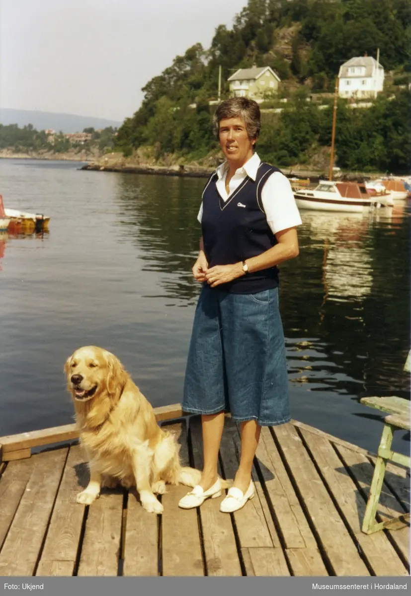 Kvinne i marineblå vest saman med hund på brygge.