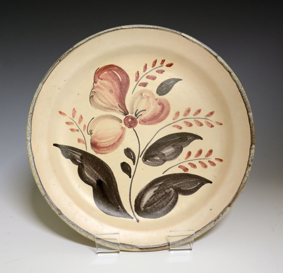 Prot: Flat tallerken av porselen med glatt kant. "Lyserødbeige" glasur. Flaten dekorert med stiliserte blomster i blågrå og brune farger. Kanten brukket opp i gråbrun farge.
