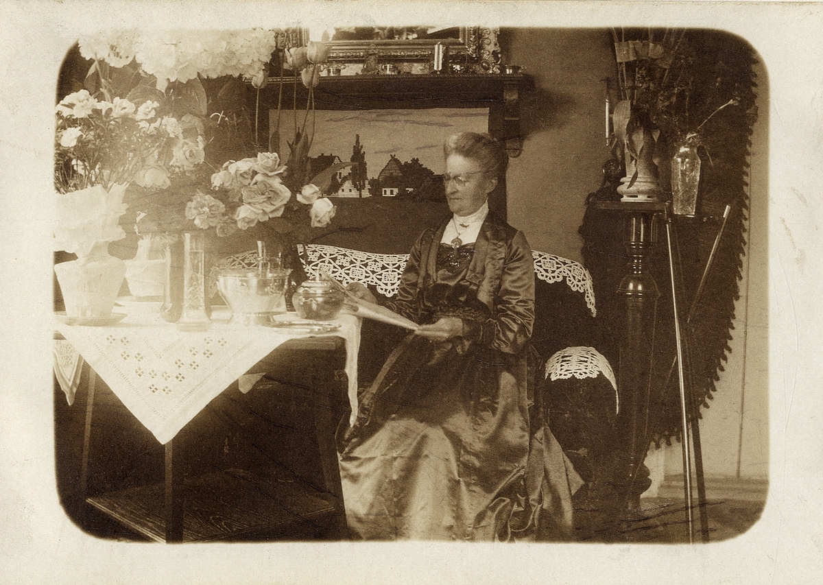 Foto av en äldre dam i lång finklänning av siden, som sitter i en soffa och läser hyllningstelegram på sin födelsedag.