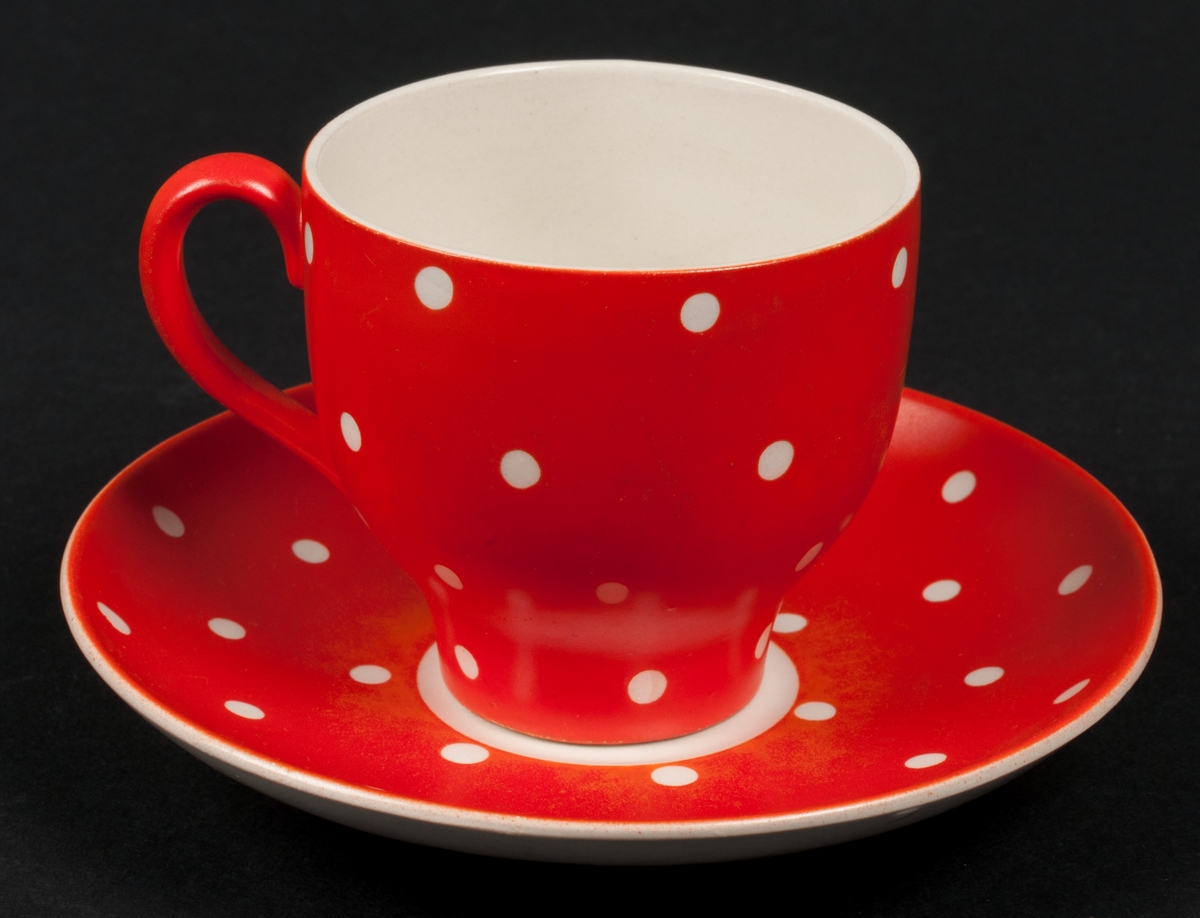 Kaffekopp (med tillhörande fat och assiett) tillverkad vid Gefle Porslinsfabrik AB. Röd bottenfärg med vita prickar. Dekorens namn "Amanita" formgiven av Helmer Ringström. Koppens modell förmodligen formgiven av Arthur Percy.