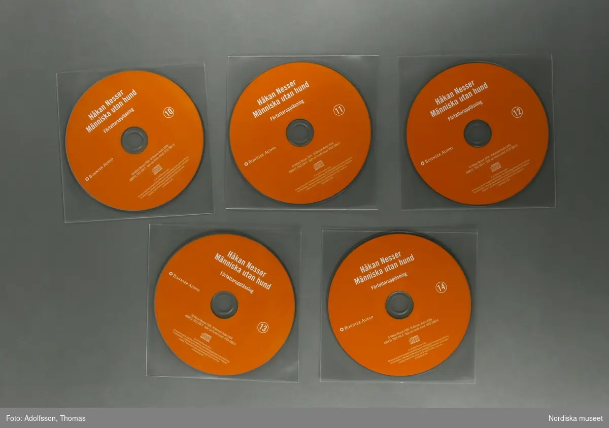 CD-bok.  a:1-2-n:1-2) 14 st CD-skivor, i en kvadratisk, transparent plastficka, innehållandes uppläsning av romanen "Människa utan hund" skriven av författaren Håkan Nesser som även är uppläsaren på denna CD-inspelning.  Inspelningen är totalt 15 timmar lång.

CD-skivorna är tunna cirkelfornade,  12 cm i diameter, på ena sidan oranga med text om titel samt kort om produktionen. Varje skiva är numrerad för att visa rangordningen i vilken skivorna ska lyssnas på/spelas. Baksidan på skivan är silverfärgad, speglliknande, och innehåller själva ljudspåren som läses av när skivan placeras i en CD-spelare.
Varje skiva är separat plackad i en kvadratisk, transparent, plastficka för att skydda mot repor som ev skulle kunna skada skivan. 
Samtliga skivor är tillsammans packade i en o) kvadratisk papperskartong/ konvolut. Konvolutet har på framsidan en bild av ett fönster med ett gultonat fotografi visandes ett fönster med en flaxande fågel utanför. På konvolutets baksida finns en introduktion till bokens handling samt  information om tillverkningen av denna CD-bok.
(CD är en förkortning av engelskans Compact Disc. CD-skivan kan lagra digital information, exempelvis musik.
CD skivans konstruktion: överst är ett transparant plastlager följt av ett tunt lager av blank metall med ett präglat spiralformat spår med mikroskopiskt små gropar som avläses av CD-spelarens laserstråle när skivan snurrar (ca 500-700 varv/minuten). Ljuset studsar tillbaka och läses som en följd av ettor och nollor, som i detta fall, blir till ljud.(Källa/. Nationalencyklopedin)


/Cecilia Wallquist 2019-02-20