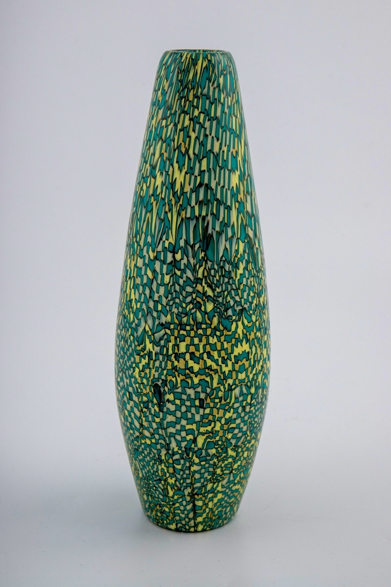 Balusterformet vase i opakt mosaikkglass, bestående av små glasstaver som skaper et geometrisk, uregelmessig mønster i hvitt, gult og turkis, omgitt av sorte konturlinjer. Sirkulær munning og bunn.