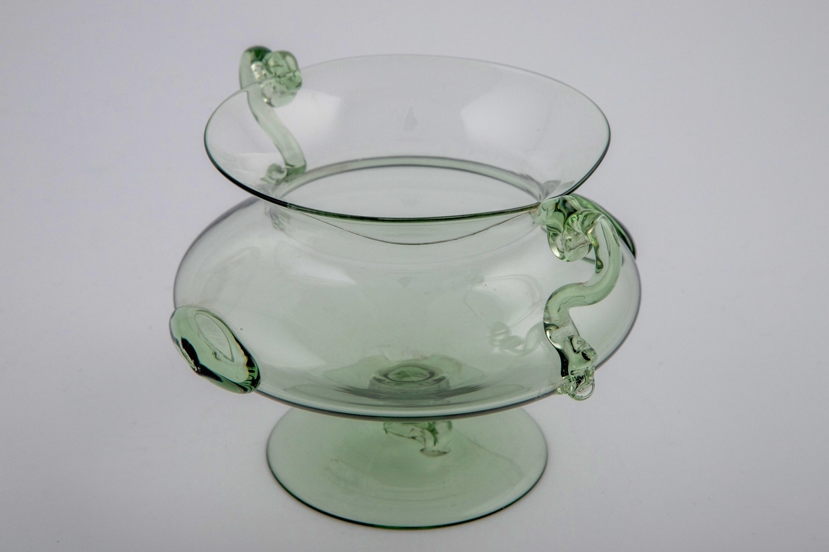 Kraterfromet vase i gjennomskinnelig grønnfarget glass. Kuleformet korpus med to ovale glassknotter, som hviler på en hevet sirkulær fot. Vasen har en lav konisk hals, og to slangeformede glasstaver fungerer som hanker.