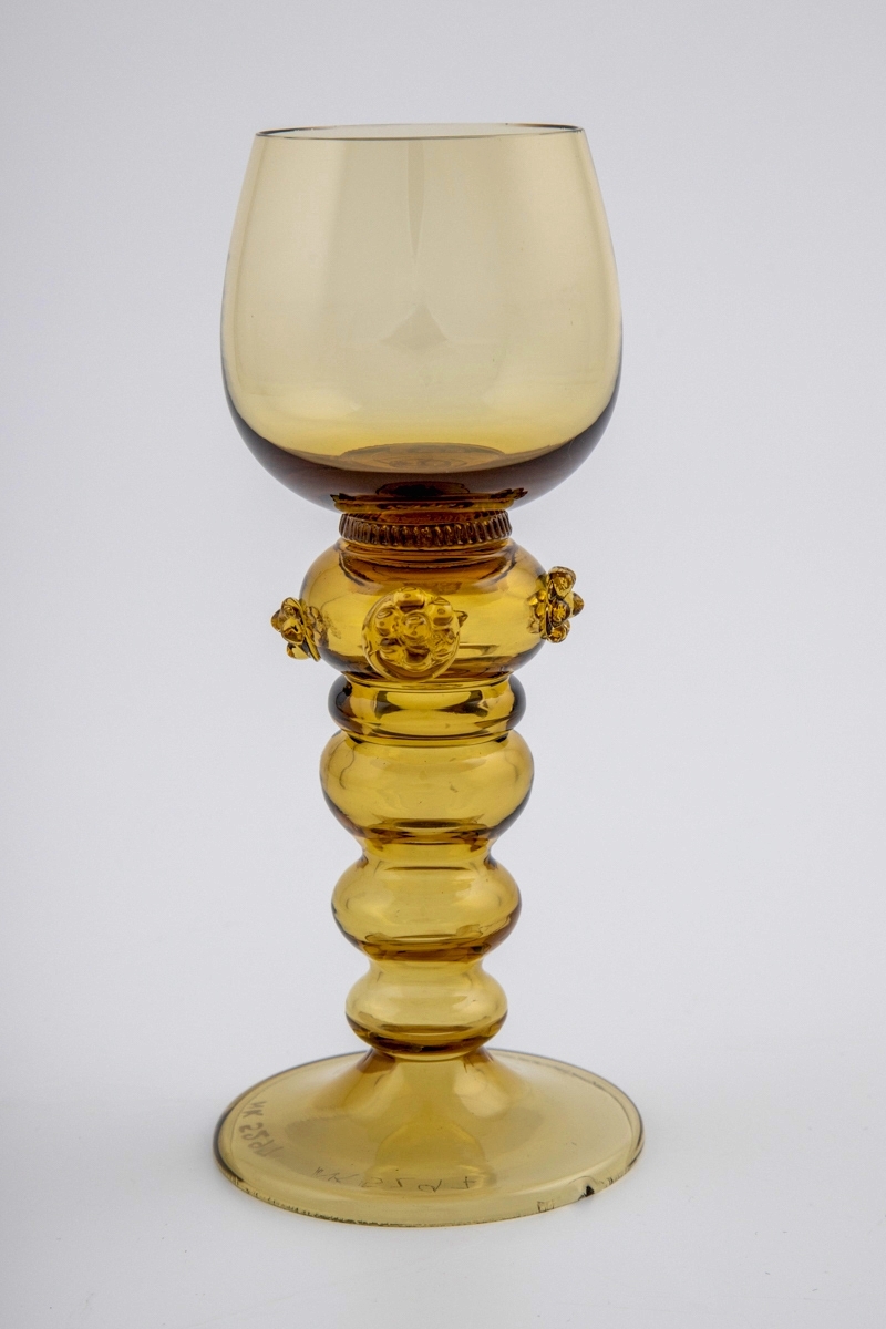 Hvitvinsglass i gjennomskinnelig gulbrunt glass. Klokkeformet kupa som bæres av en konisk stett bestående av ringformede vulster, hvorav den øverste er dekorert med tre rosetter ("bringebær"). Sirkulær fotplate som er noe hevet.