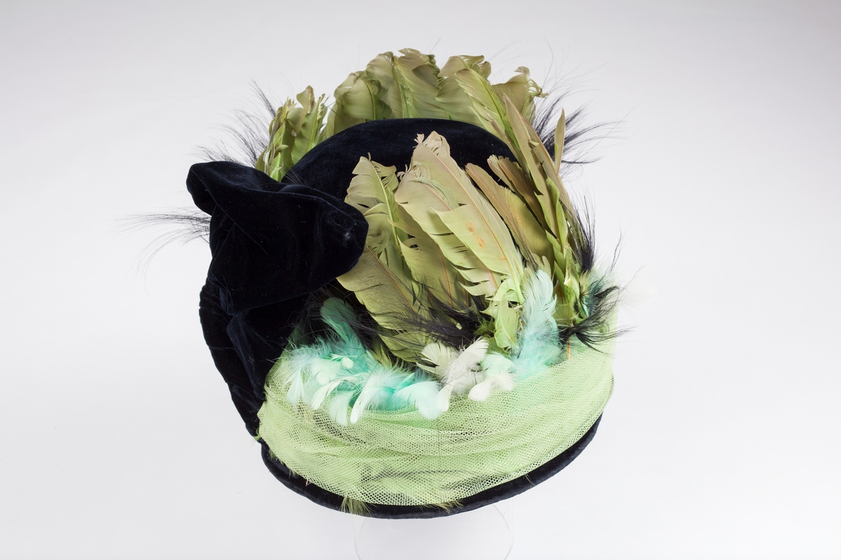 Svart fløyels hatt med dekor av fjær og tyll i grønt.