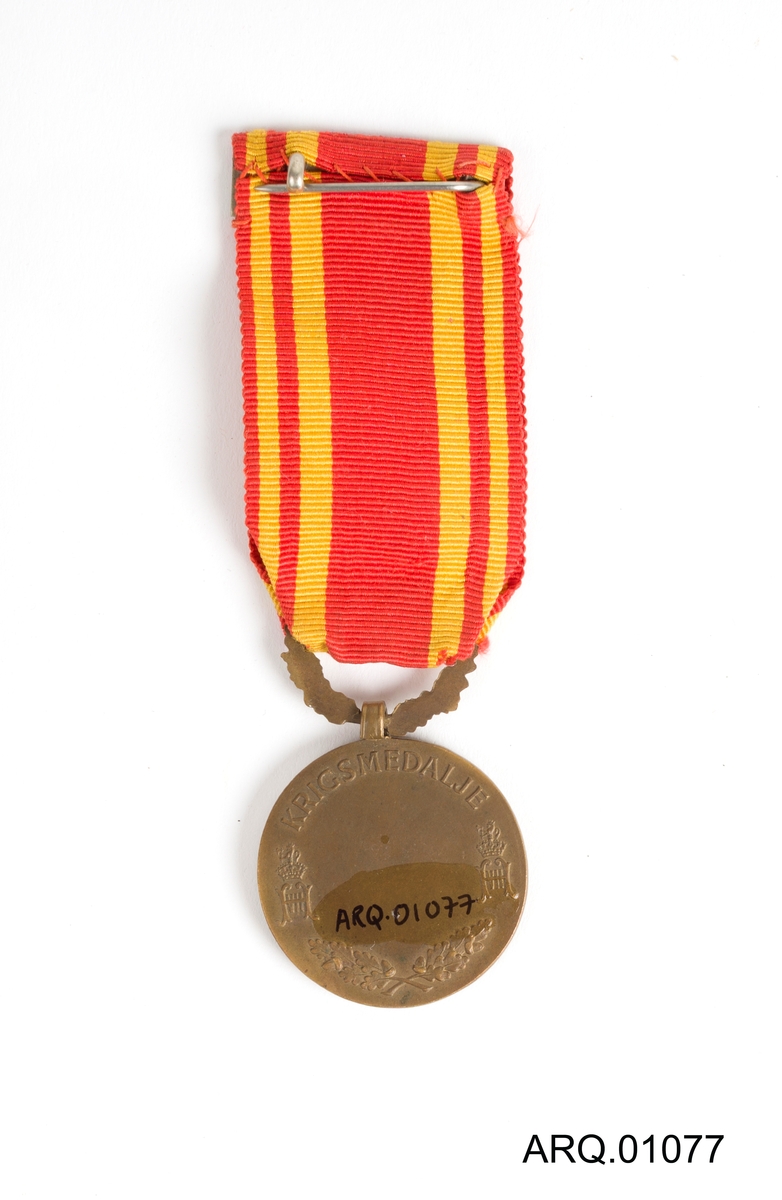 Medalje med Kong Haakon VII på. Tekstilet er rødt med gule loddrette striper. Festenål på baksiden av tekstilet.