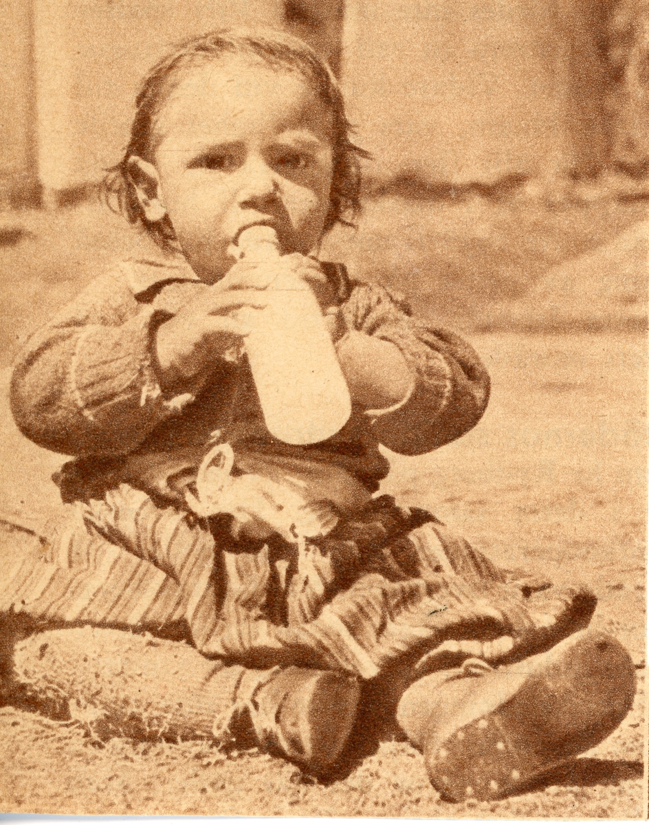 Ett barn sitter på marken och dricker ur en flaska. Bildens ursprung är okänt.