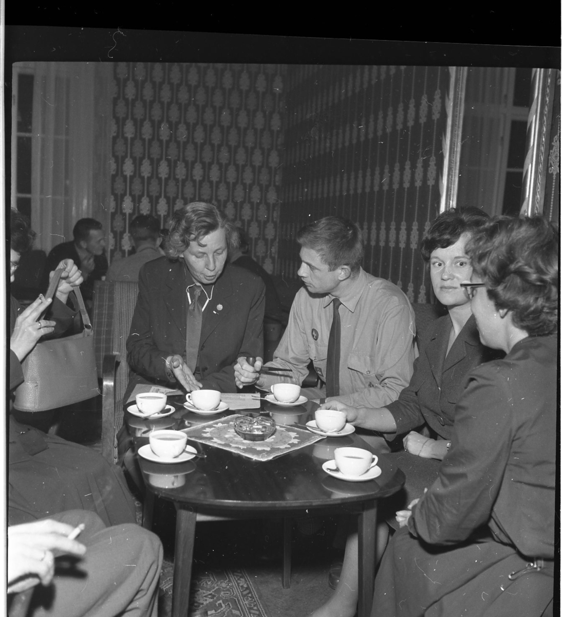 Män och kvinnor, en del i scoutdräkt, sitter i grupper runt bord med kaffekoppar och cigaretter.