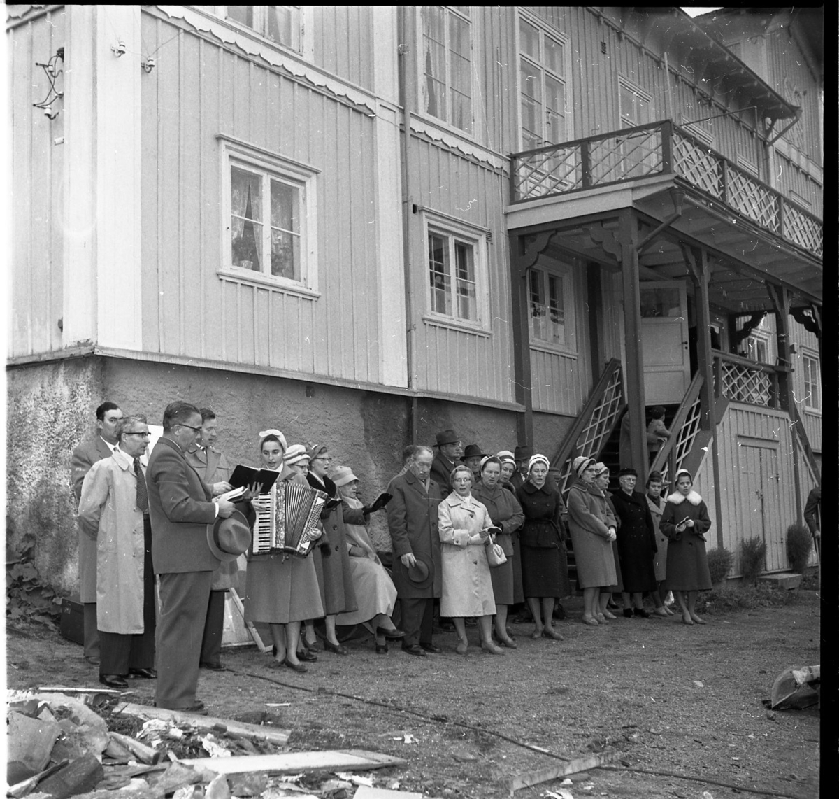 En grupp män och kvinnor på baksidan av Brahegatan 75a (Hjertströmska gården) står och sjunger till ackompanjemang av en kvinna som spelar dragspel. Närmast i bild står pastor Robert Svensson