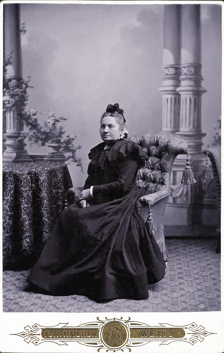 Foto av en äldre kvinna i mörk, höghalsad klänning och s.k. frumössa. Hon sitter i en "emma" vid ett bord med lång duk. 
Helfigur. Ateljéfoto.