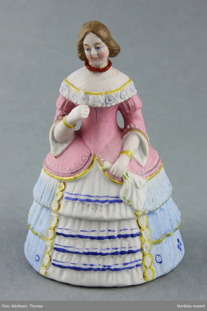 Huvudliggaren:
"Ask med lock, porslin, parian; locket utformat som dam iförd krinolin; brunt hår, rosa liv, ljusblå kjol med infällning fram i vitt med mörkblå ränder, grön rosett bak; bårder i guld och guld; rött halsband; näsduk i ena handen; möjl. porträtt av Jenny Lind (1820-1887). Enl. uppgift tillhört Jenny Lind."
