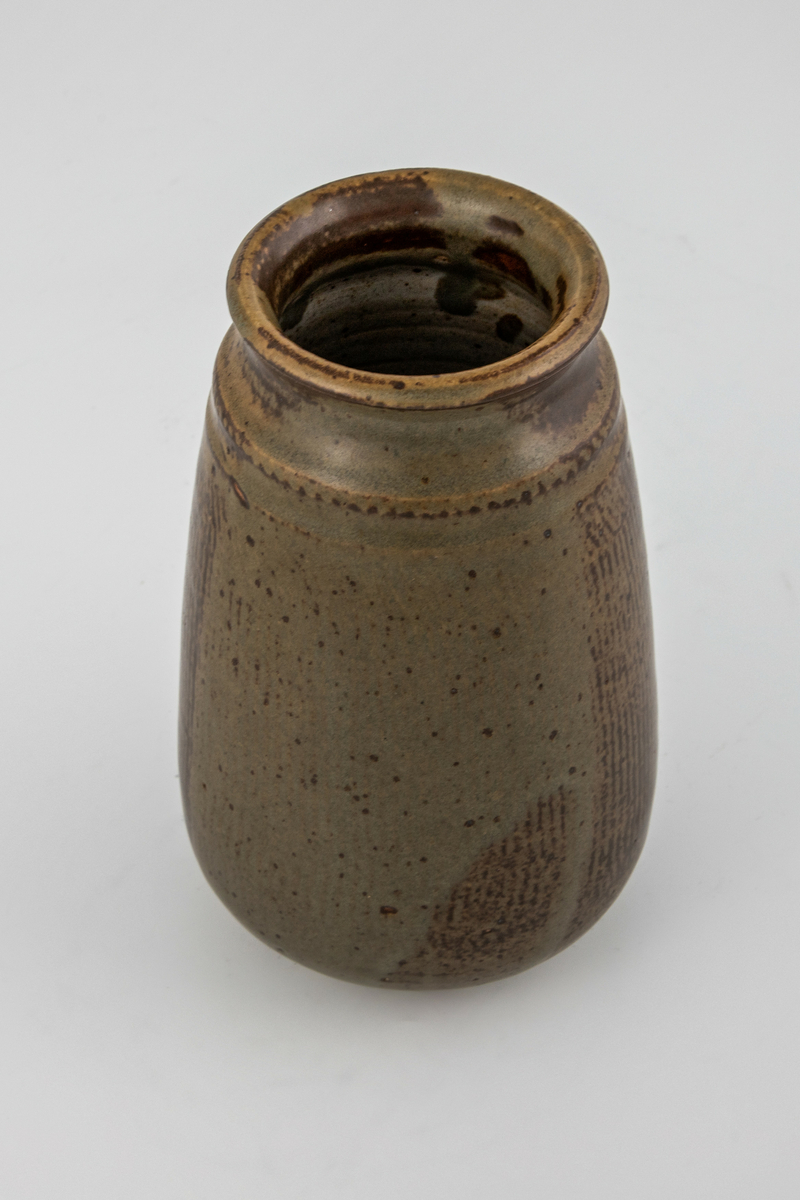 Høyreist vase i glasert steingods. Kjegleformet korpus som hviler på en lav fotring, utoverbrettet munningsrand. Korpus er dekorert med vertikale mørkebrune riller, som på deler av vasen er dekket av en gråbeige flyteglasur.