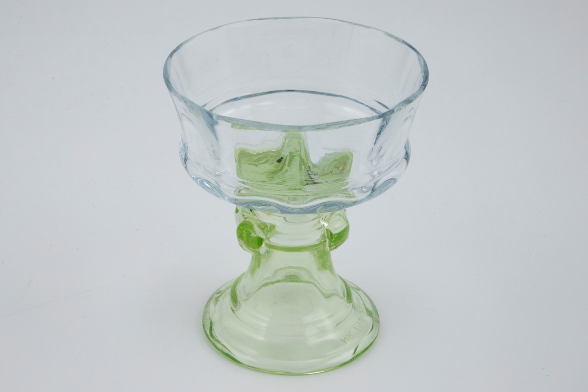 Begerformet drikkeglass. Kupa er svakt rillet og utført i klart glass. Glassets kraftige fot er lysegrønn, derav verkets tittel pied vert (fr. grønn fot). Nederst på foten er det innrisset: "Cristallerie Gallé à Nancy".