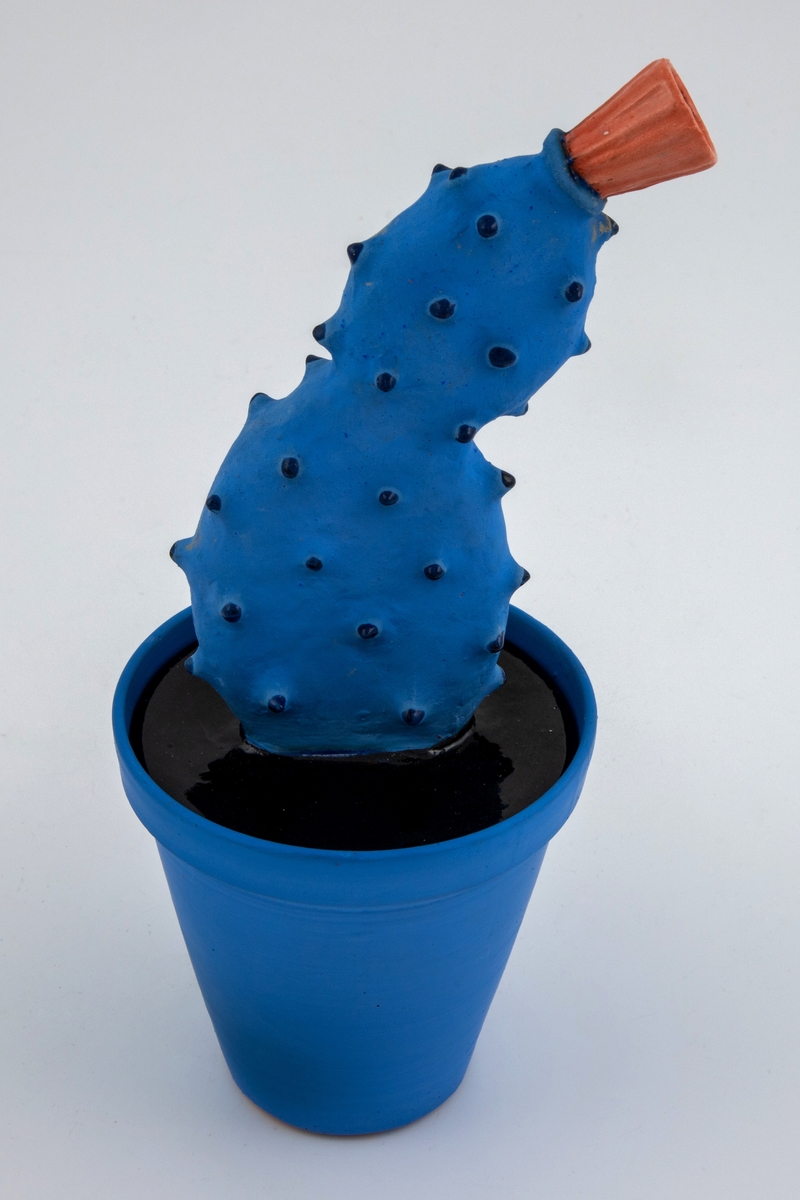 Blå blomsterpotte med skulpturelt lokk i blått, svart og oransje. Bunnens form er en rettavkortet kjegle med utvidelse øverst, der lokket hviler nedi sporet 1,4 cm fra øvre kant. Til det flate lokket sitter en kaktusform som danner et forvokst håndtak. Kaktusen hviler organisk til den ene siden, har utstående, avrundede pigger og en stilisert blomst på toppen.