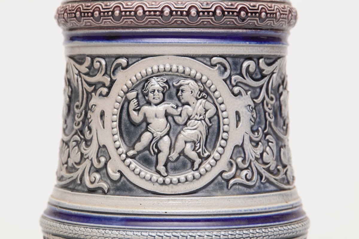 Blått, grått og fiolett leirkrus med lokk av tinn, profiler øverst og nederst. Dekor: relieff, ranke, bukkehoder og to dansende figurer.