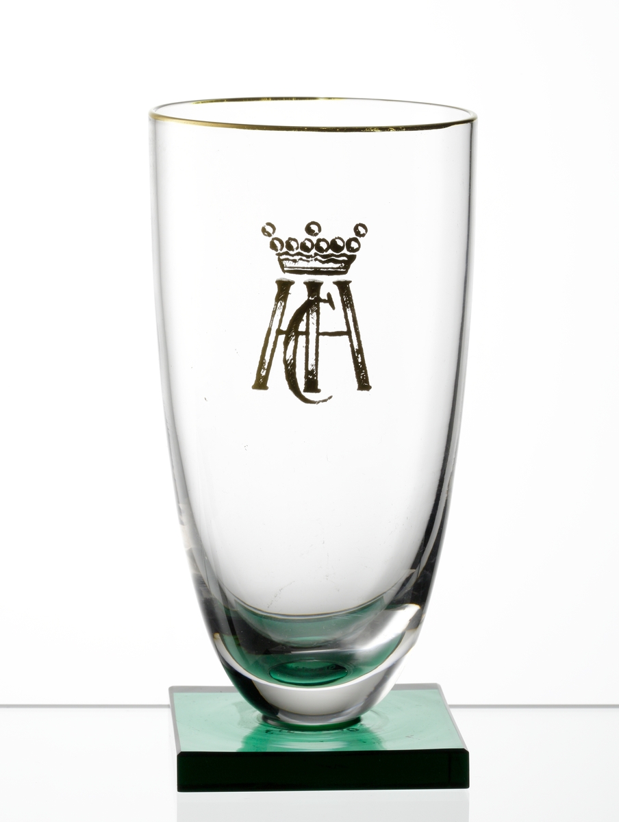 Champagneglas med slät kupa och grön, slipad, fyrkantig fot. Guldmynning, guldmonogram (under friherrlig krona).
Formgivare: Simon Gate.