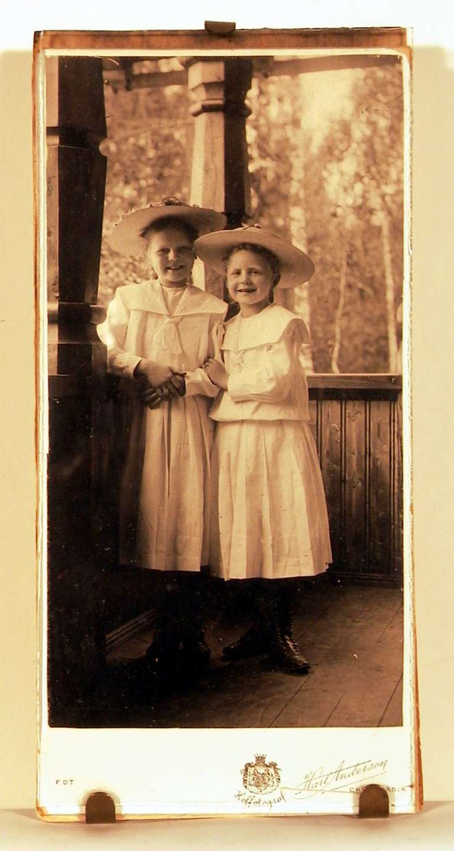 Barneportrett; to store piker med støvler og hatt - med stor skygge og dekorasjon rundt hattepullen. Pikene står på en overdekket veranda med naturen som bakgrunn.