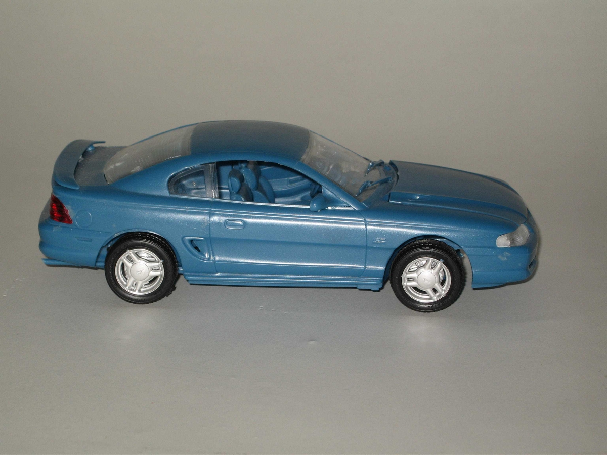 Leketøysbil som modellbil satt sammen av mange deler, hvorav karosseri og understell er laget av blå plast, vinduer og framlykter av klar plast, baklykter av rød plast, dekkene av svarte plast og felgene av kromfarget plast. Bilen er en modell av en Ford Mustang.