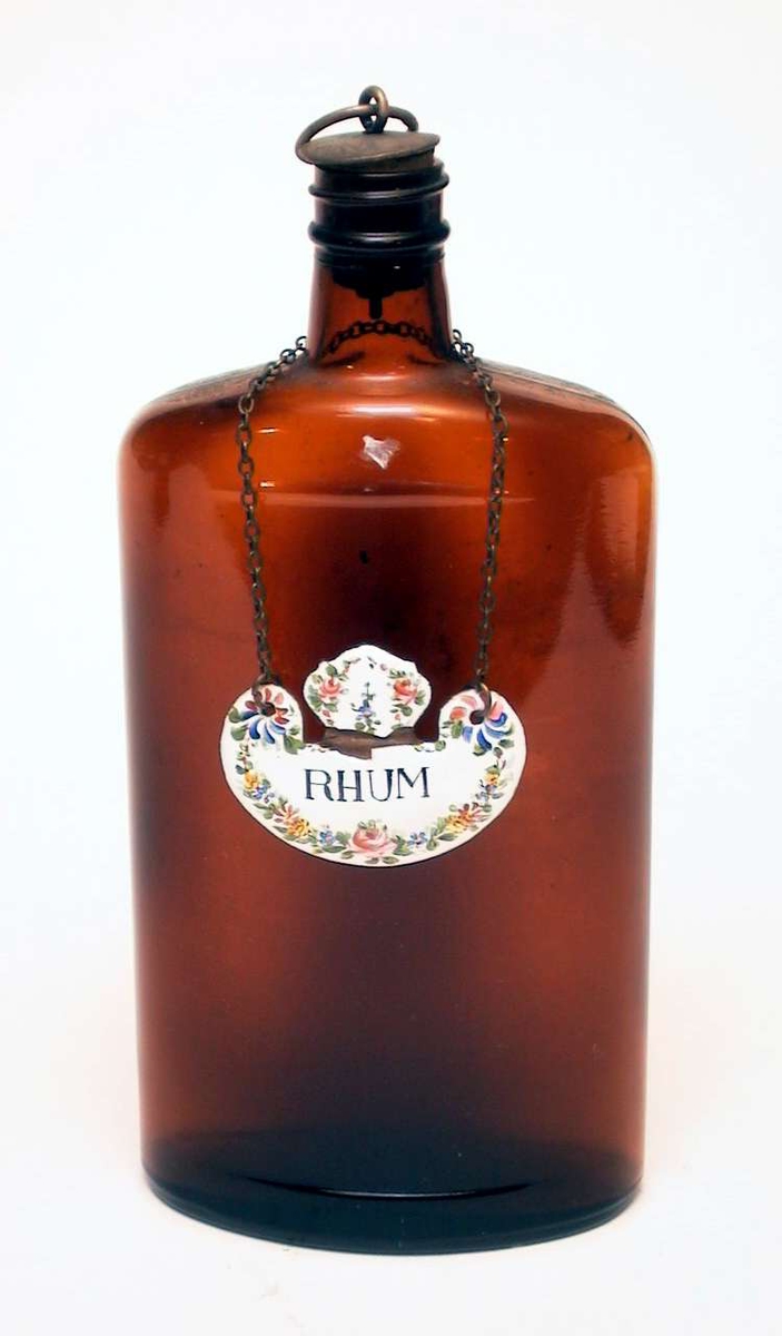 Flat brennevinsflaske i brunt glass med kork. Korken har plate og ring av metall på toppen. Rundt halsen på flasken henger et metallskilt i lenke. Det er dekorert med blomster og er påskrevet 'Rhum'.