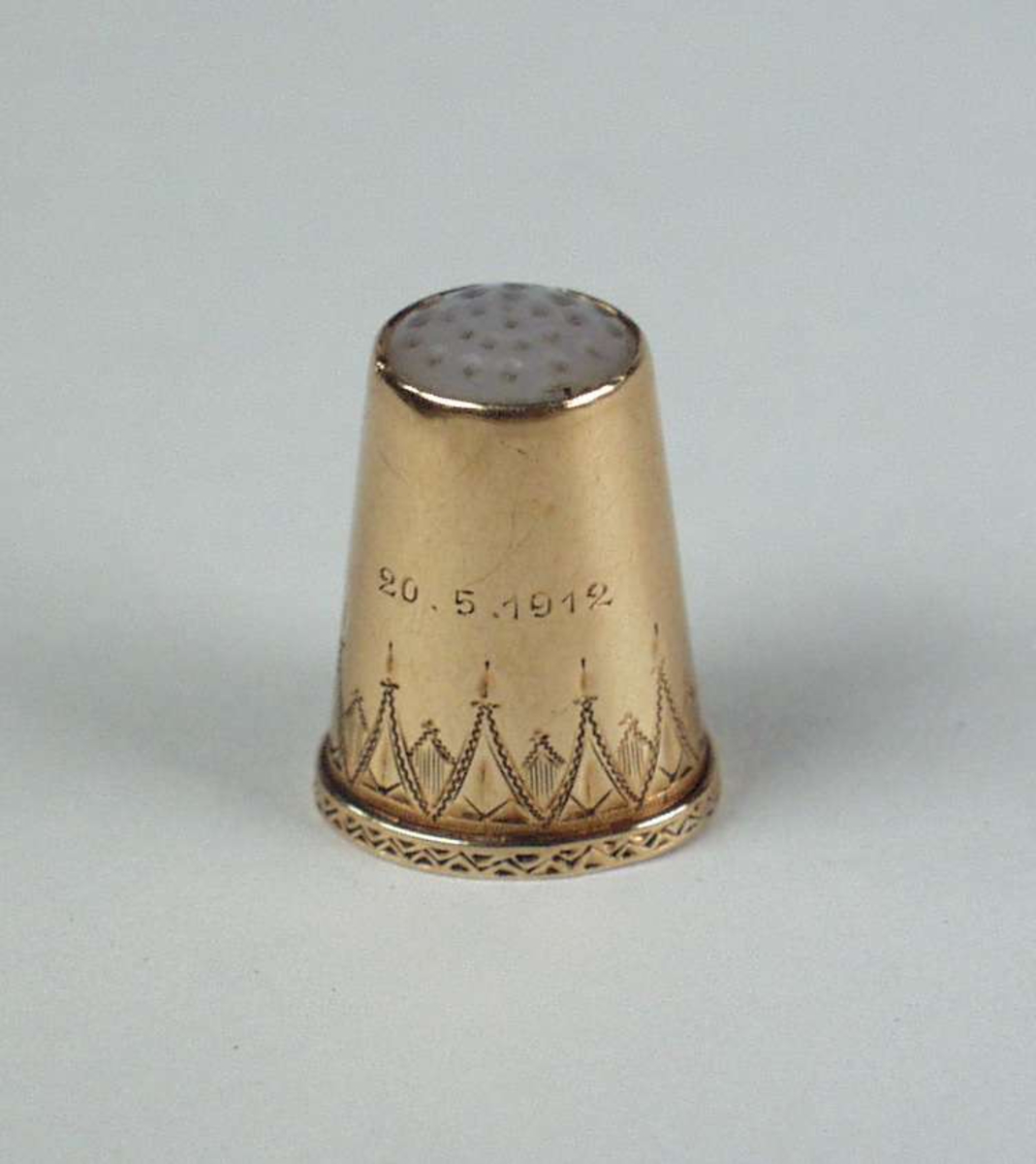 Fingerbøl av gull med topp av perlemor. SUs initialer og datoen 20.5.1912 er inngravert. Fingerbølet har gravert dekor.