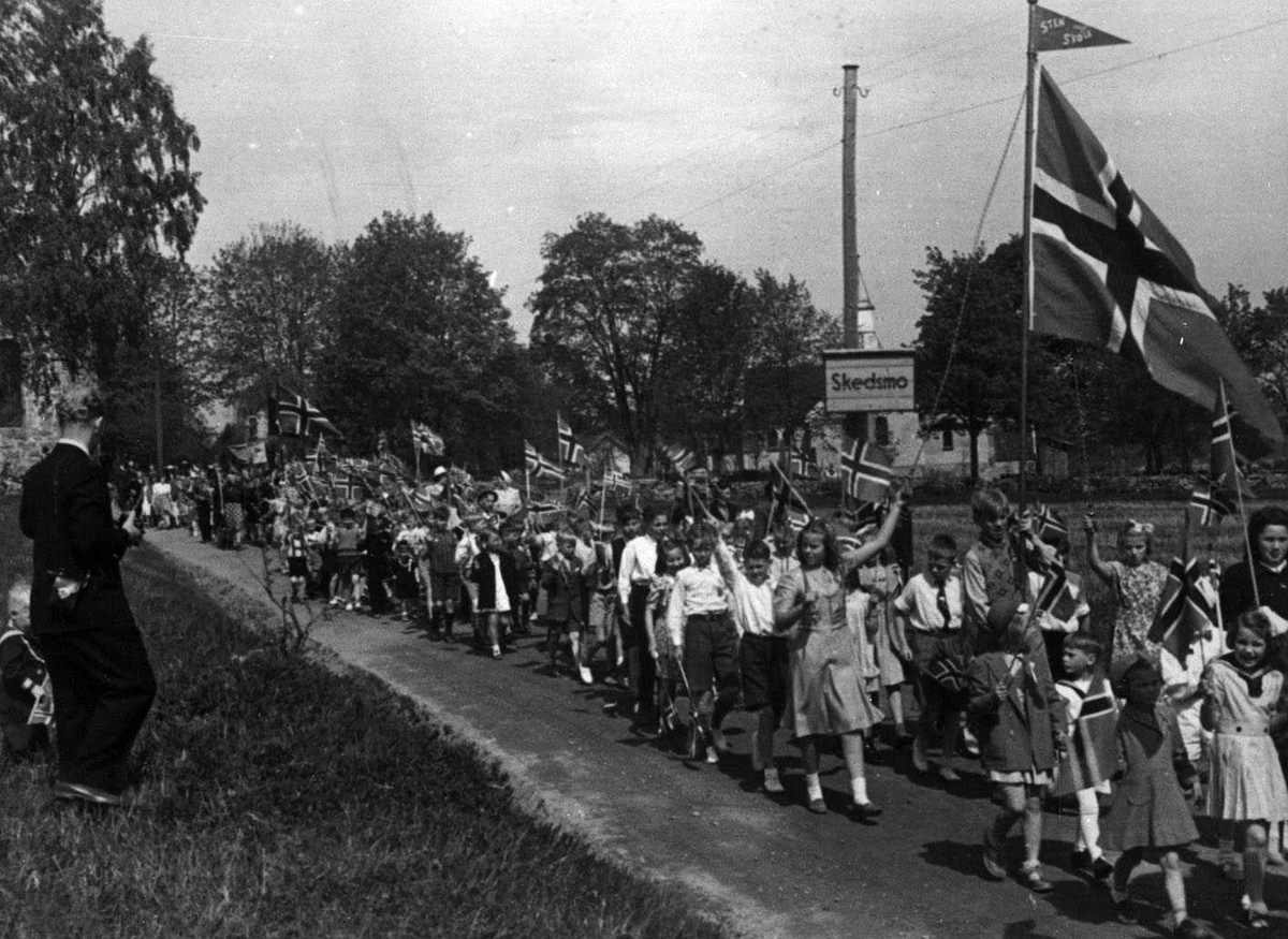 17. mai feiring ved Skedsmo kirke 1945. 17.maitog på marsj. Barnetog På toppen av hovedflagget en vimpel med teksten Sten skole.