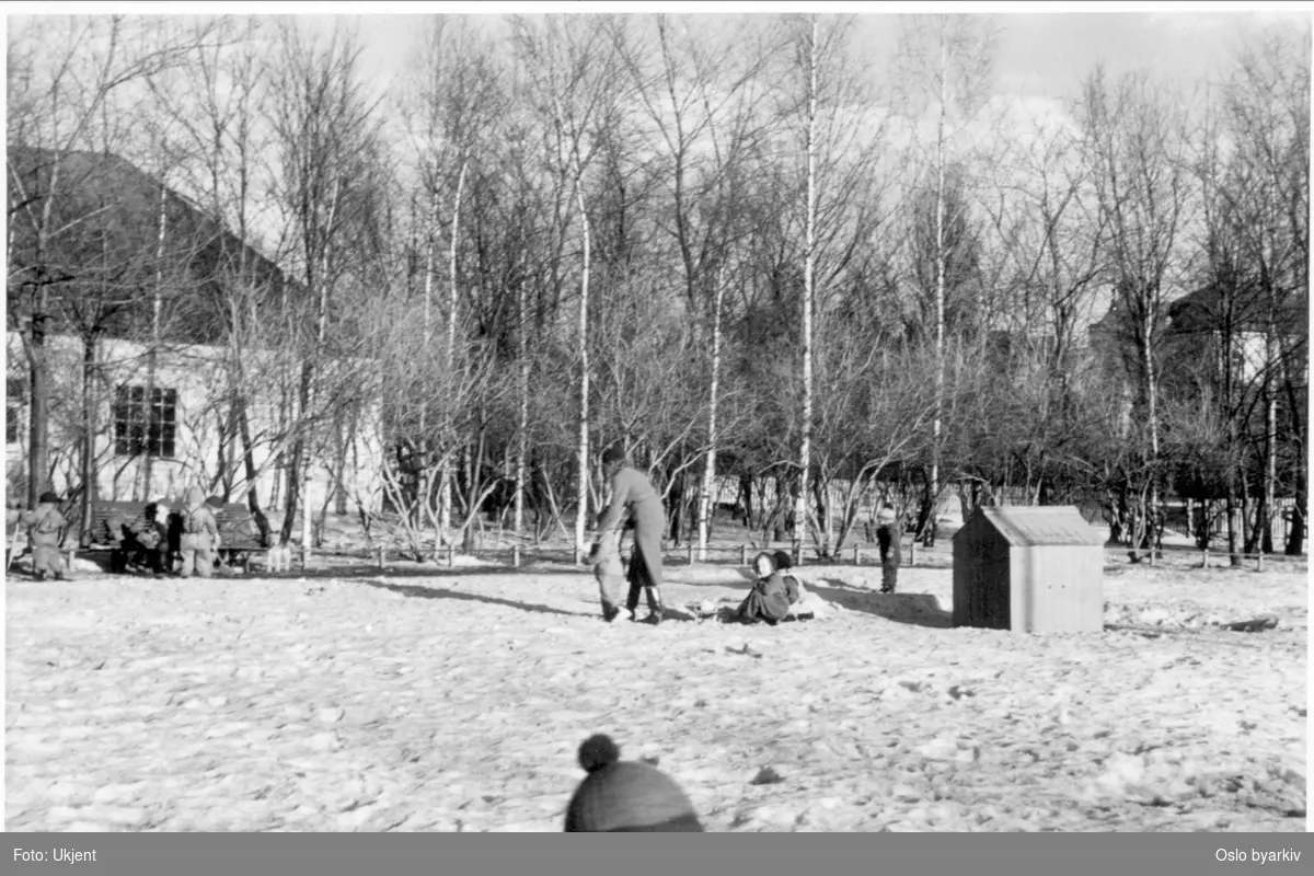 Vinter i Frognerparken. Lekende barn på skarasnø ved lekeplassen sør for hovedinngangen til Vigelandsparken.Sørøstlige del av parken. Hvitt stakittgjerde langs Kirkeveien i bakgrunnen.