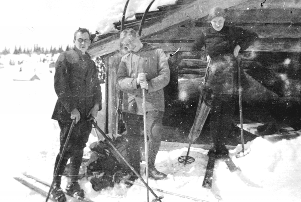 Tre personer på ski foran "Hotel Mikkelsrud", laftet tømmerhytte på Ringsakerfjellet. Vinter, fritid, påske.