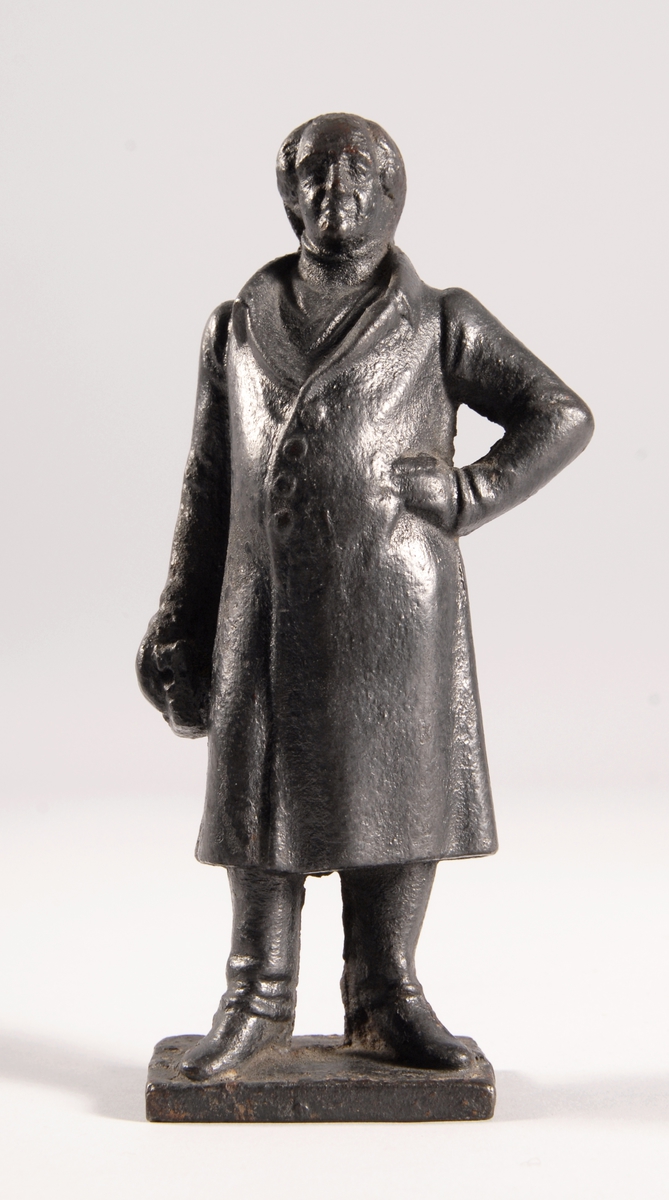 Liten statuett støpt i jern som forestiller enstående mann med frakk.