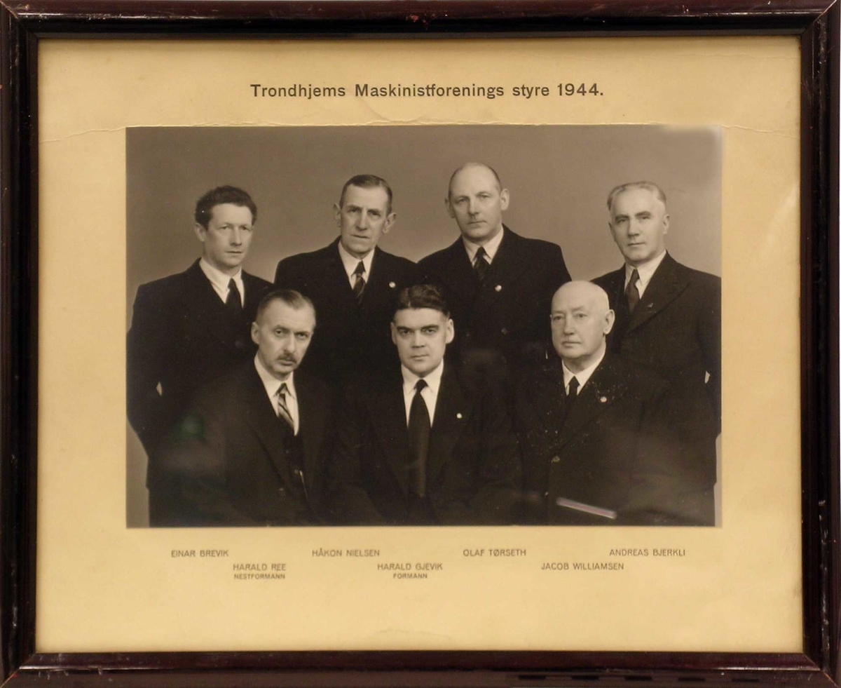 Gruppefotografi av 7 av medlemmene i styret for Trondhjems Maskinistforening i dress tatt i et atelier.