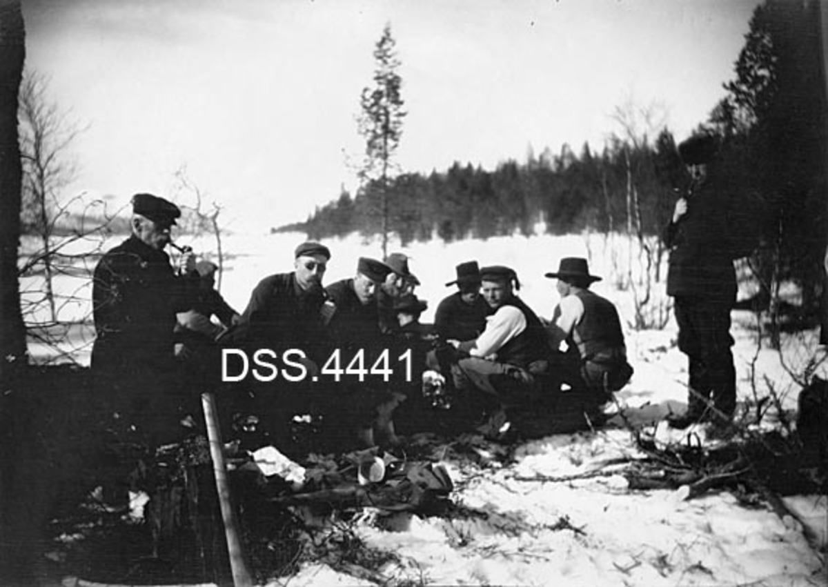 Åtte mannfolk, samlet til pause i forbindelse med tømmermerkingsarbeid ved Stenvika på Femund.  En mann står (til høyre), de andre sitter.  Flere av karene røyker pipe. 