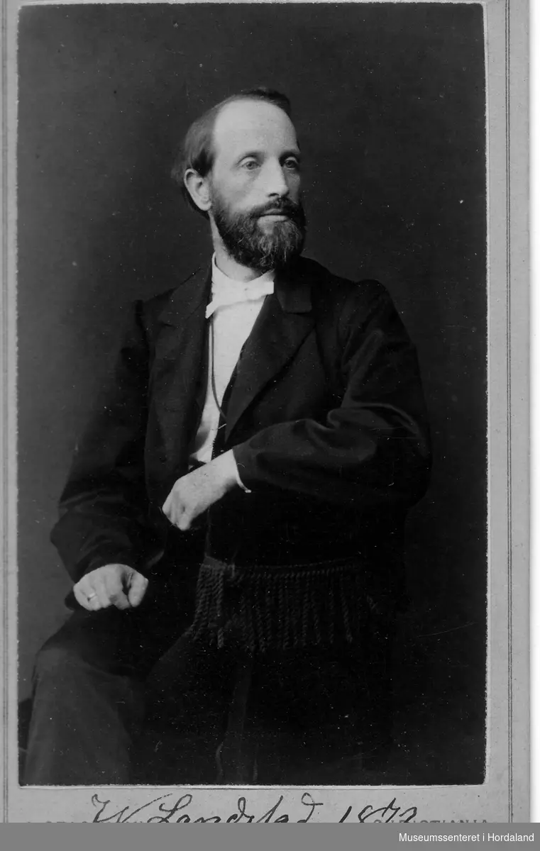Portrett av mann med bart og skjegg i lenestol, vendt mot venstre (høgre på foto), eine handen over armlenet, andre handen knytta i fanget.