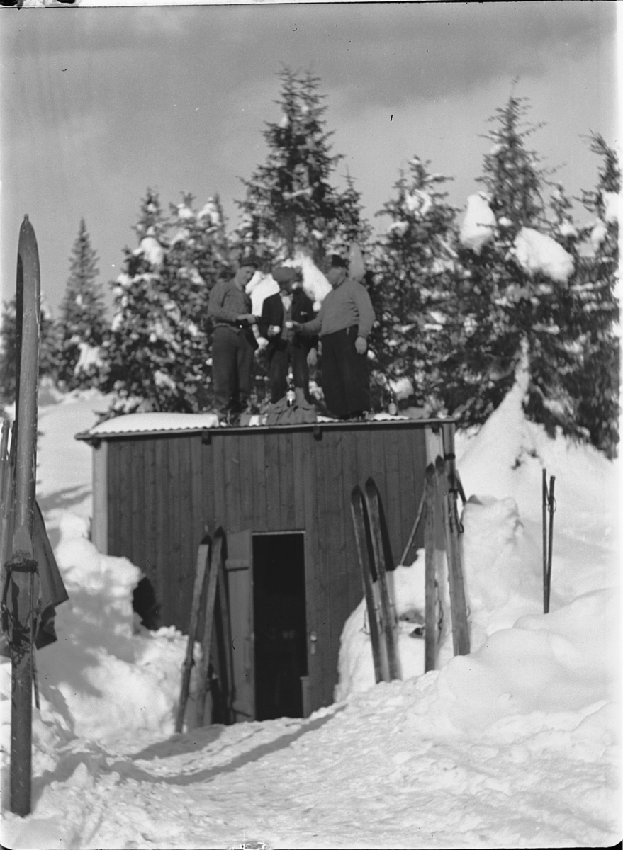 Tre menn på skitur tar seg litt drikke på taket av ei hytte. 