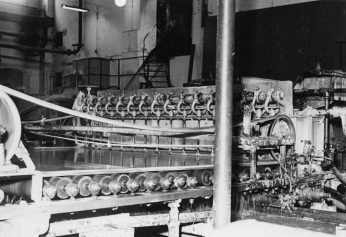 Innløpskasse og tilstøtende deler av wirepartiet på PM 4 ved Drammenselvens Papirfabrikker på Modum i Buskerud.  Herfra ble silt papirmasse ledet inn på sjølve papirmaskinen.  Denne komponentens utforming var viktig for å sørge for at massen ble jevnt fordelt både på tvers av og langs banen, slik at papirstrukturen ble jevn. Fotografiet ble tatt i 1953. 