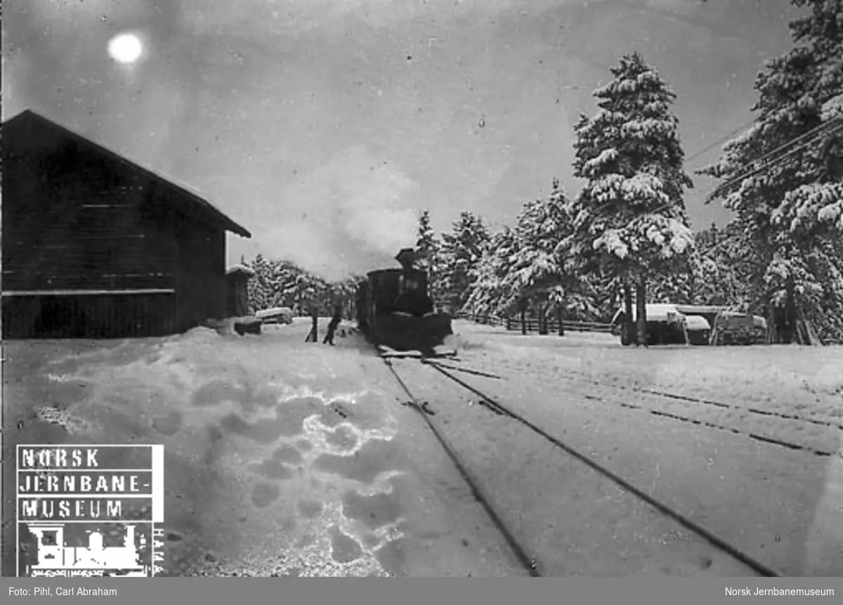 Vinterbilde med damplokomotiv type VII med stor frontplog som ankommer med tog, muligens på Rena stasjon