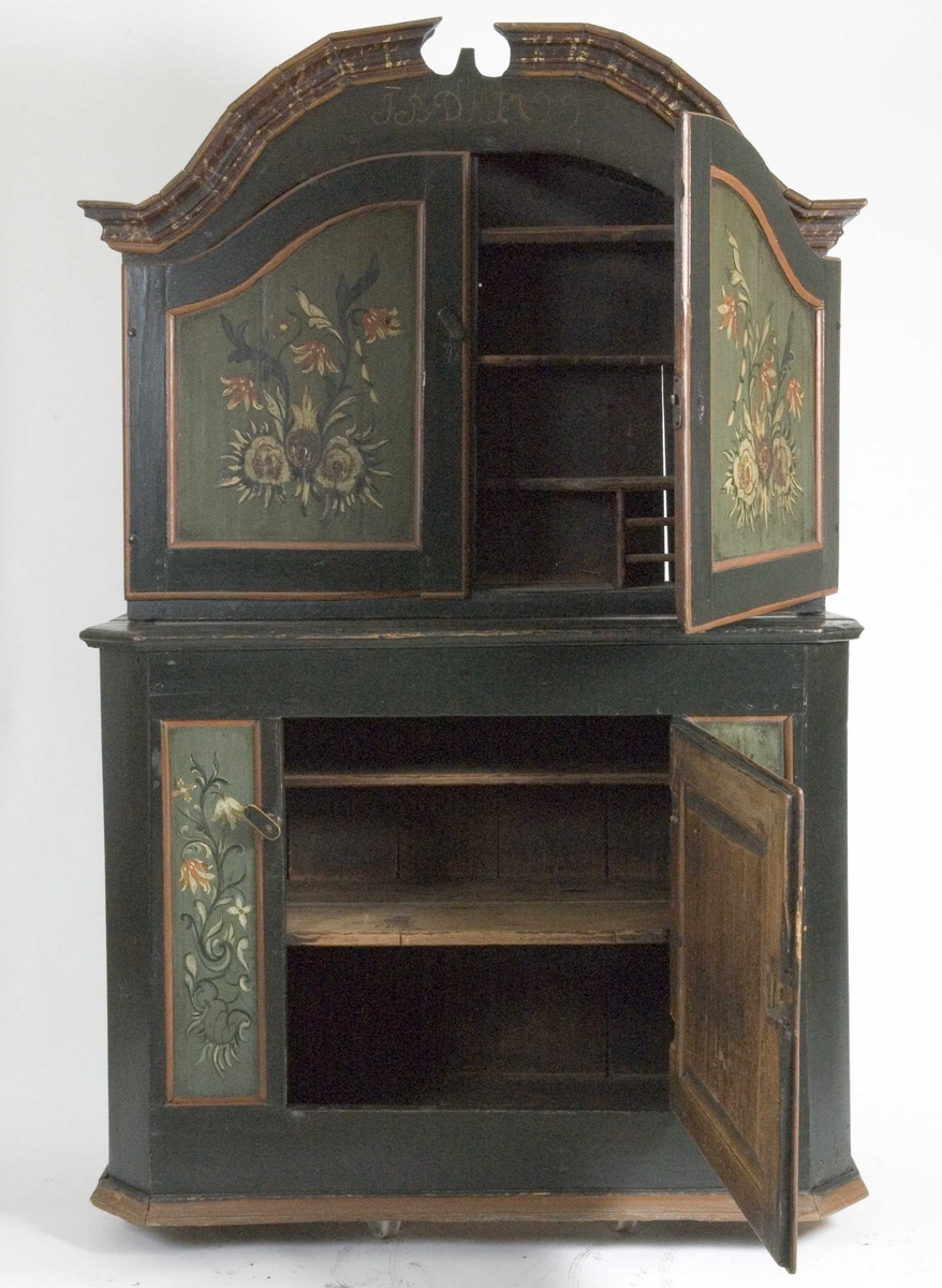 Skap i over- og underdel. Ovedelen har skjehylle og plass til skuffer. Buet topplist satt sammen av vinkelskårete deler. Blomstermalt.