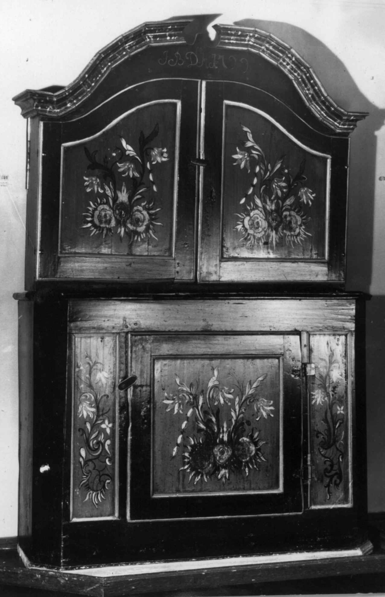 Skap i over- og underdel. Ovedelen har skjehylle og plass til skuffer. Buet topplist satt sammen av vinkelskårete deler. Blomstermalt.