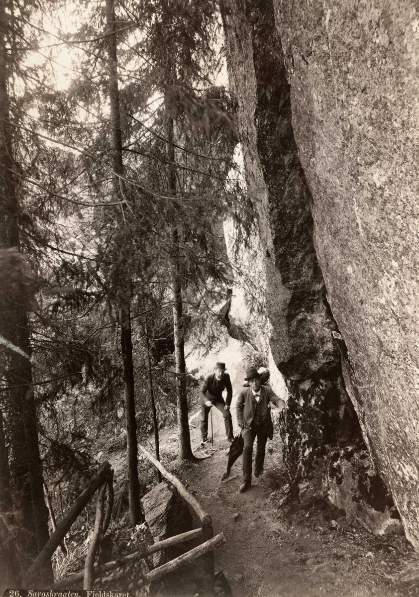 Sarabråten, Østmarka, Oslo. To menn spaserer på vei i skogen, under klippe.