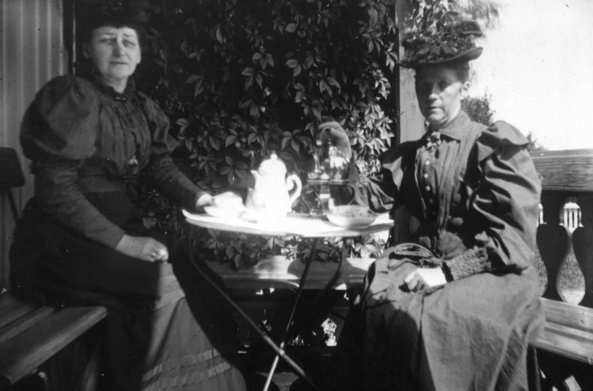 Div. drakt, Norge 1908-10. To kvinner ved et bord på en veranda. Den ene i lang kjole og med hatt. Den andre i kjole og skjørt.  