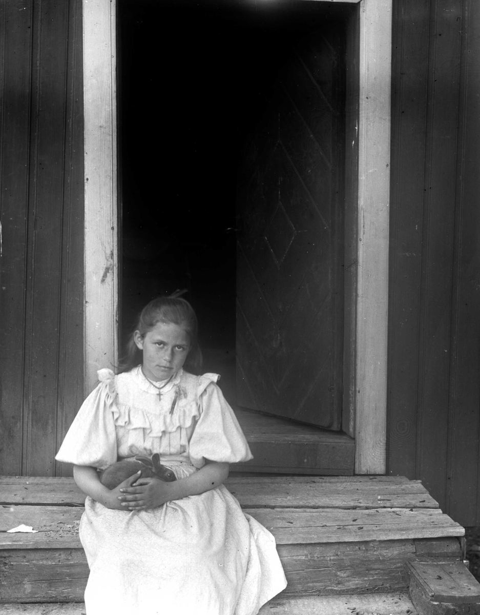 Portrett av jente, Emily Holm (f.1886), fotografens niese, sittende på en trapp foran en halvåpen dør, kledd i lys sommerkjole. Hun har en kanin i fanget. Muligens tatt på kapellangården Bergenhus i Rakkestad  (til 1897)eller prestegården i Hurum (fra 1897) , der faren var prest. 
Fra malerinne Juliane Fredrikke Langbergs (1856-1930) samling, tatt ca.1895-1910 med motivkrets slekt og venner samt steder tilknyttet samvær med disse.