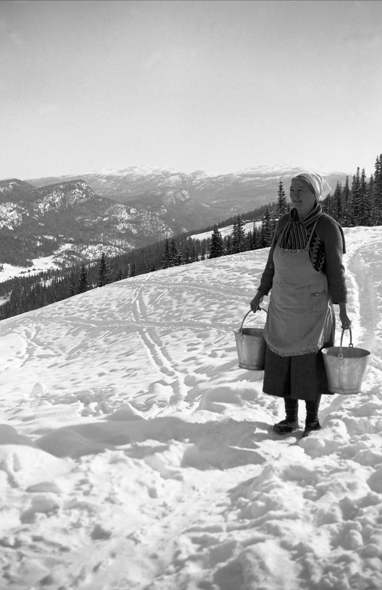 Hushjelpen Rakel Hansen bærer to bøtter med vann i et snøkledd fjell. Fotografert påsken 1940.