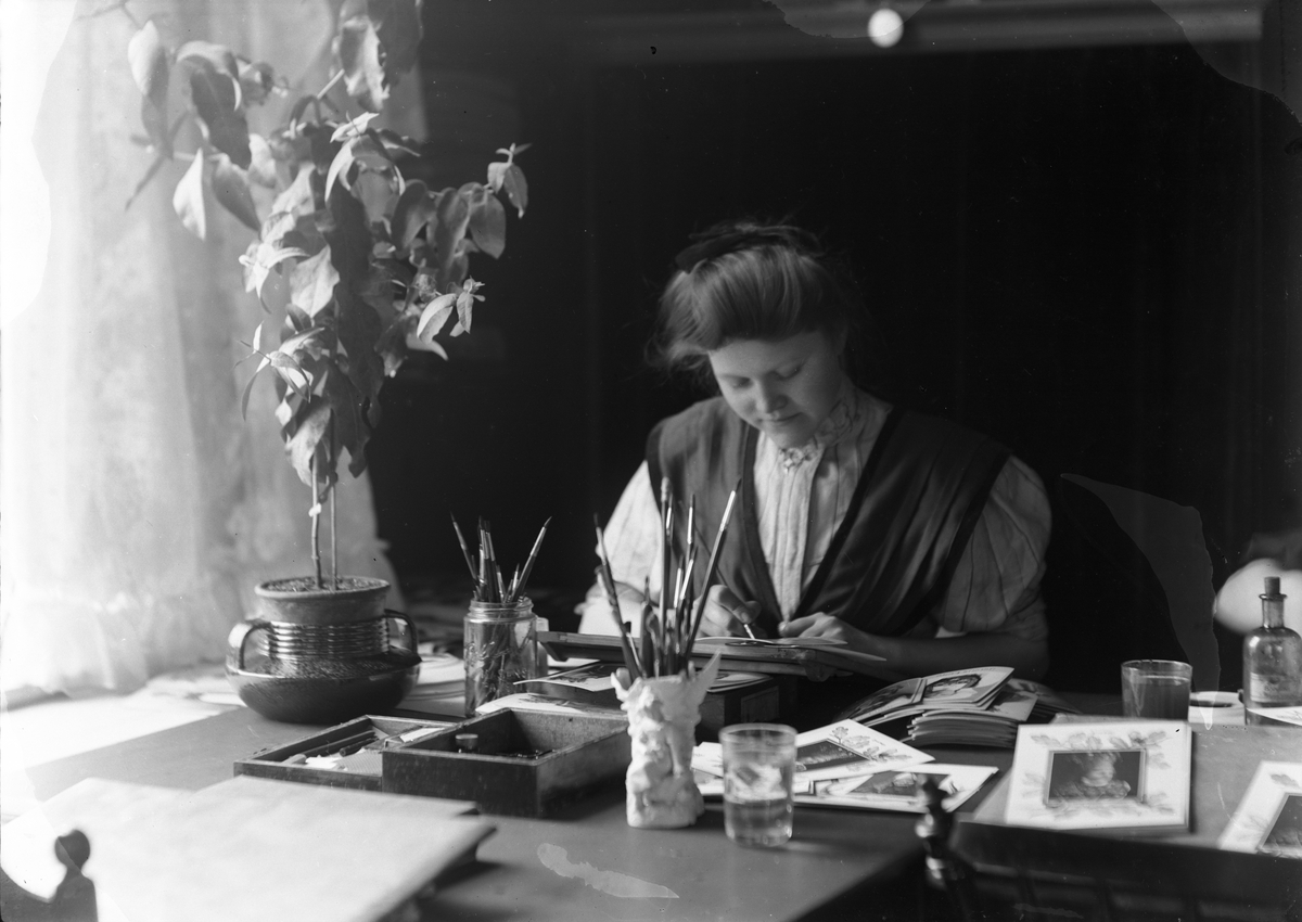 Antakelig Solveig Lund selv i studioet sitt arbeidene med kolorering av papirfotografier. Fotografier og pensler på pulten.