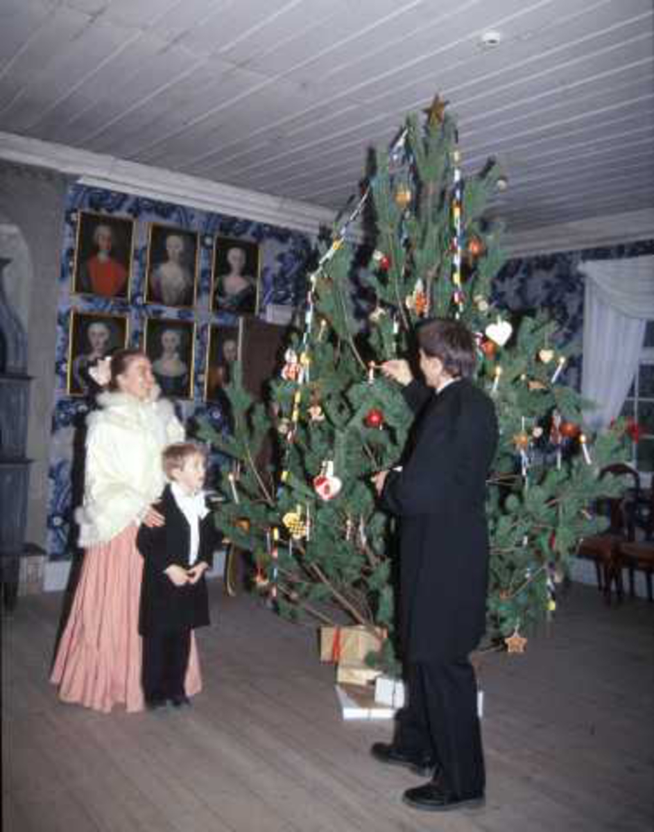Det feires jul i Leikanger prestegård, bygning nummer 191 på Norsk Folkemuseum.Museumsansatte  samt et barn, alle i kostymer er i ferd med å tenne lysene på juletreet.