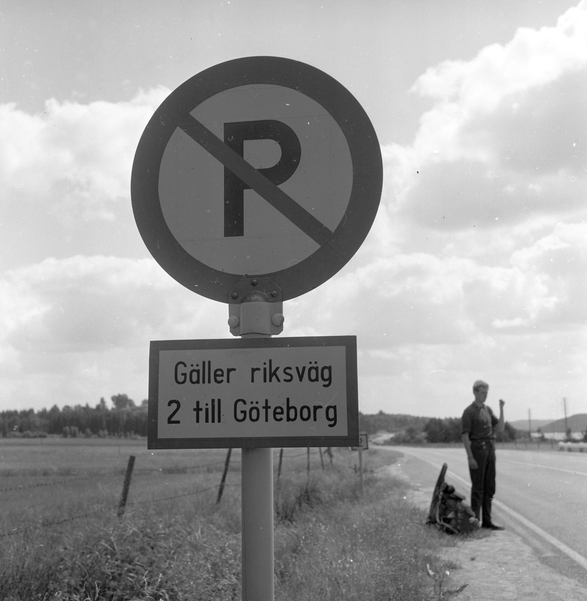 Langs Riksvei 1 står det en mann og haiker og et P-skilet med "Gäller riksväg 2 till Göteborg". Fotografert 7. juli 1958.