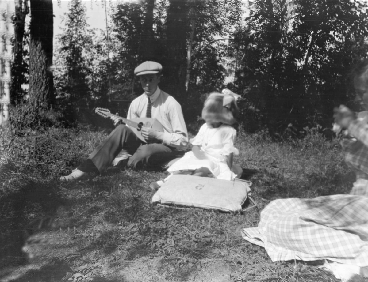 En mann sitter å spiller mandolin for en jente i skogen. Robsahm og Lund.