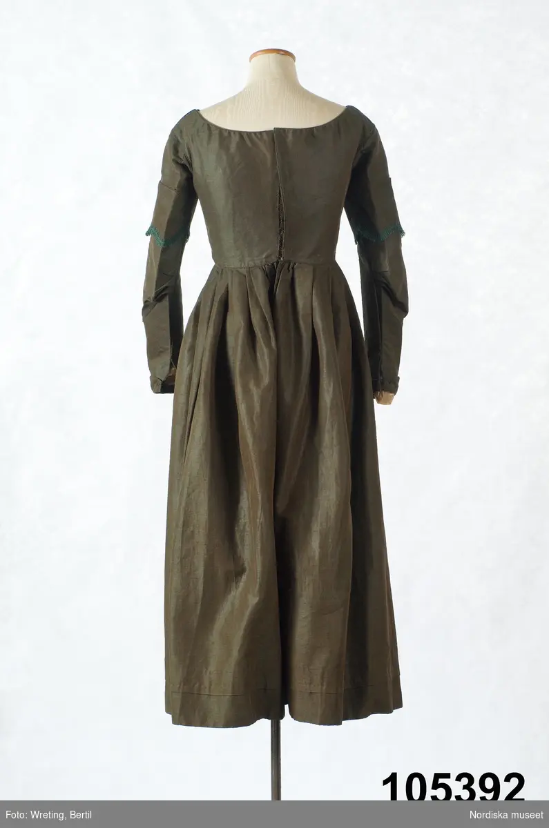 Hel klänning i 1830-talets modesnitt, av fint halvylletyg i kypert med varp av ljusbrunt bomullsgarn och inslag av grönt tunt glansigt redgarn som ger en grönskimrande yta. Livet går ned nästan till midjehöjd, figursytt med 2 framstycken med bakåtflyttade sidsömmar, ett ryggstycke med 6 inprovningar fördelade i två grupper. Vid båtringad halsringning, knäppning med tätt sittande hakar och hyskor. Isydda långa ärmar svängda med 2 sömmar  med 3 veck vid armbågen. Ärmsprund knäppt med hyska och hake samt 4 cm uppslag med rundade hörn. Upptill på ärmen besättning av tyget med stora rundade uddar kantade med klargrön bomullsfrans.  Passepoaler av tyget i alla sömmar. Livet fodrat med skotskrutig bomullslärft i rött, grönt, gult och vitt och i ärmarna ett smalrandigt bomullstyg i blått och rosa. Kjolen sydd av  5 våder, lagda veck runtom i midjan. sprund mitt fram. Uppsytt veck  en decimeter från fållen. Skoning med brun bomullslärft. Handsydd.
Har burits 1838 som ungmorsklänning, alltså av bruden dagen efter bröllopet, av Maria Margareta Mårtensdotter från Kysings, Vall sn på Gotland. Möjligen tillsammans med sidenhalsklädet 105385.
Anm. I  gott skick.
OBS Klänningen sitter bakochfram på fotona, knäppningen är fram.
/Berit Eldvik 2011-06-13