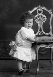 Portrett av Laila Aavatsmark i kjole ved stol. Piken er ober