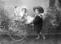 Gruppe, gutt i matrosdress med stråhatt holder barnevogn med