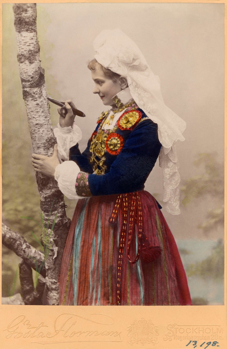 Flicka klädd i dräkt från Herrestad eller Järrestad i Skåne står och ristar i ett träd.