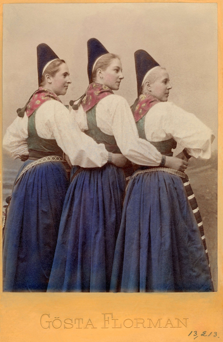 Porträtt av tre unga flickor i likadana folkdräkter från Rättvik i Dalarna. Kolorerat foto.